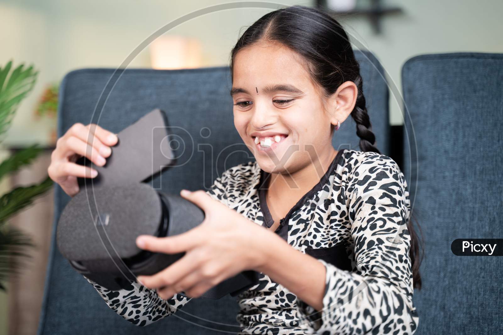Happy Smiling Young Girl Kid Placing Mobile Phone Inside The Vr Or Virtual Reality Goggles While Sitting On Sofa And Trying To Use It At Home.