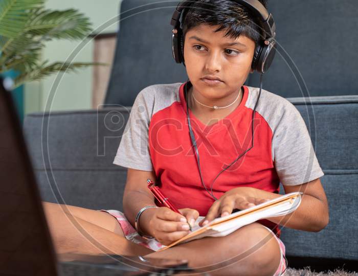 Serious Kid With Headphone Noting Down To Book By Seeing Laptop During Online Class At Home - Concept Of Online Classroom, Online Education, Technology And Lifestyle.