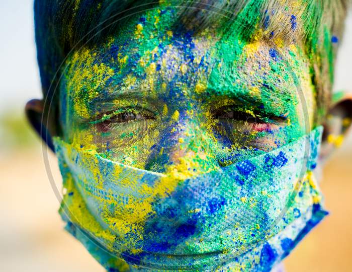 Head Shot Of Young Boy With Medical Face Mask Played Holi The Festival Of Colours And Looking Into Camera - Concept Of Holi Celebration During Covid-19 Coronavirus Pandemic.