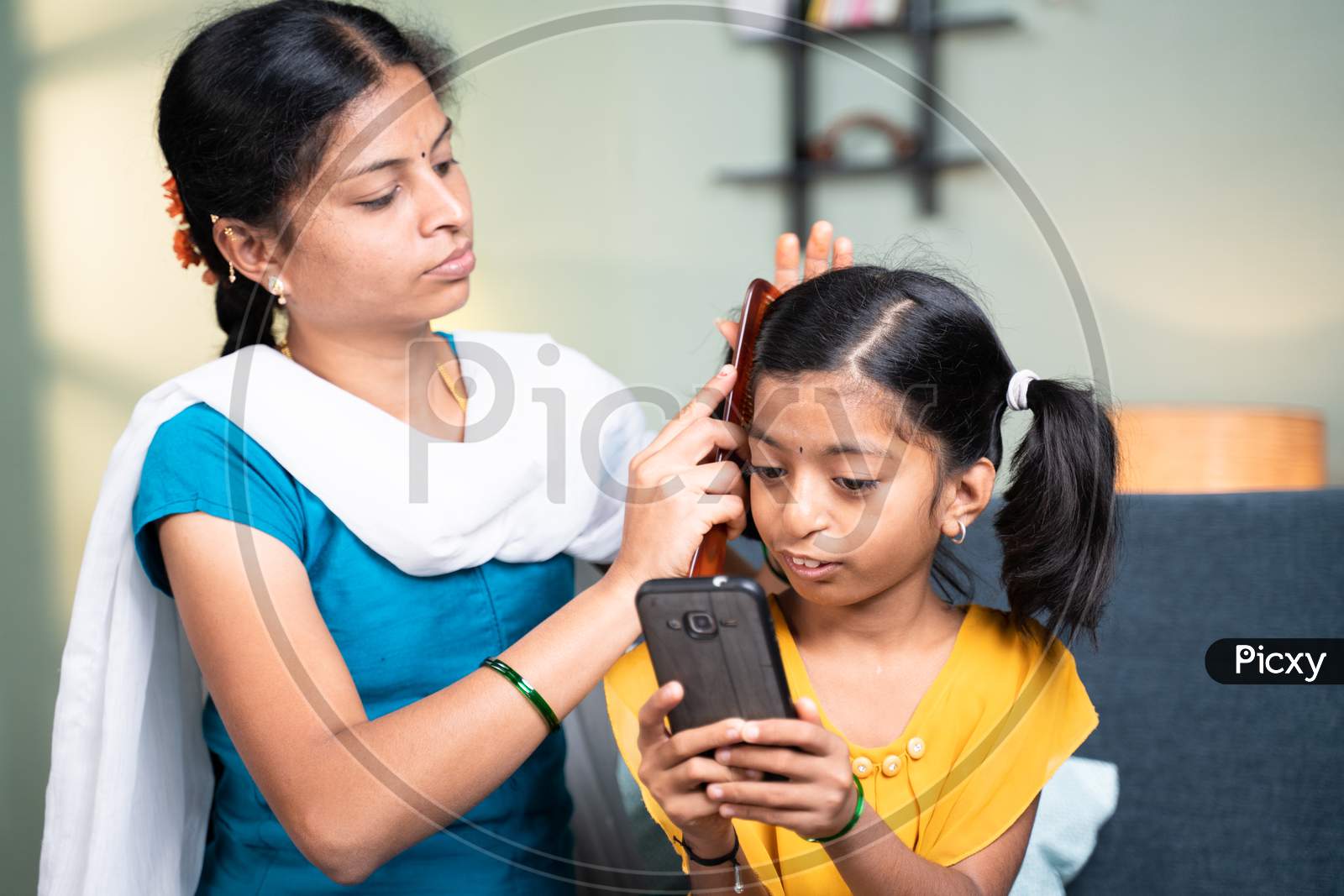 Mother Combing Her Kid Hair While Kid Busy Using Mobile Home While Sitting On Sofa At Home By Ignoring Mother - Concept Of Kids Mobile Phone, Game And Technology Addiction.