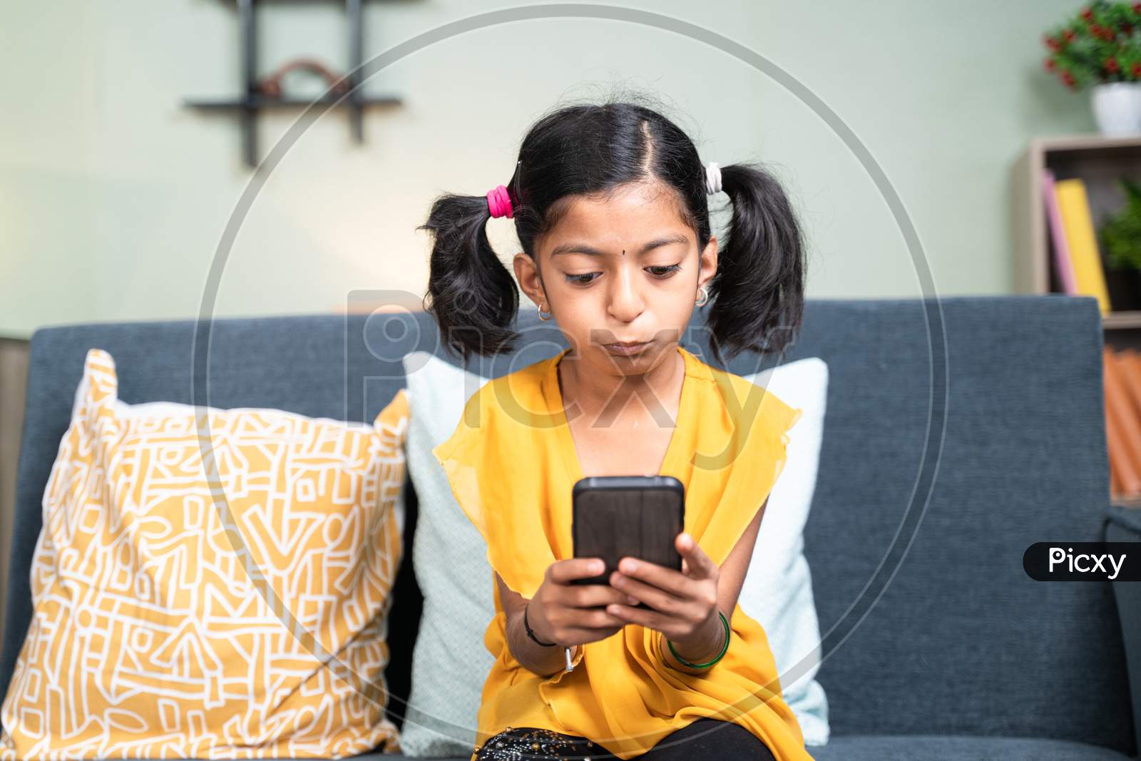 Young Girl Kid Using Mobile Phone While Sitting On Sofa In Bending Head Posture - Concept Of Kid Mobile Phone Game Addiction, Technology And Modern Lifestyle.