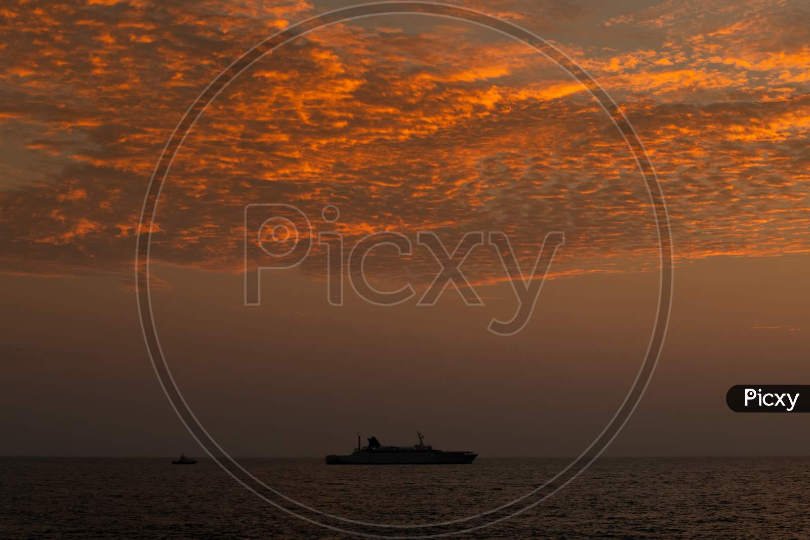 Silhouetted Of A Long Distanced Ship In Horizon Of The Ocean, After The Sunset Golden Hour Photograph, Clouds Glowing In The Evening Sunlight.