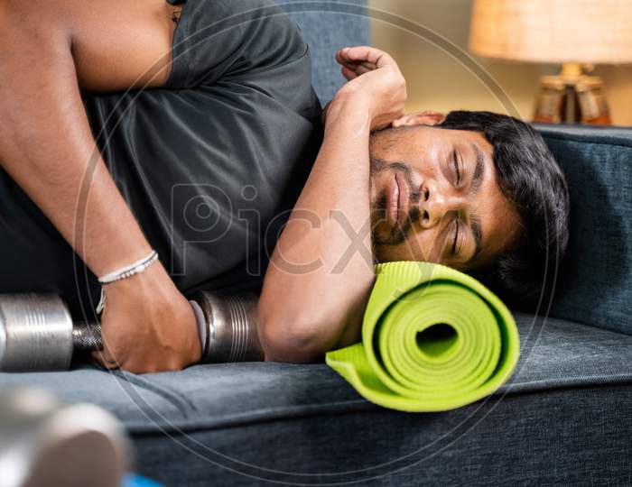 Tired Young Man Sleeping On Sofa By Holding Dumbbells And Yoga Mat - Concept Of Healthcare, Lifestyle And Fitness Man Resting After Workout In Home Gym.