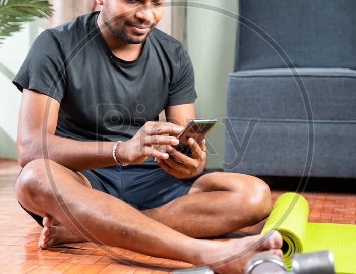 Young Man Busy Using Mobile Phone During Work Out - Millennial Checking Online Exercise Tutorials For Workout - Concept Of Home Gym Due To Coronavirus Covid-19 Pandemic.