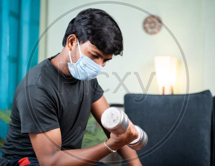 Young Man With Face Mask Busy In Work Out Or Doing Exercise Using Dumbbell At Home - Concept Of Home Gym Due To Coronavirus Covid-19 Pandemic