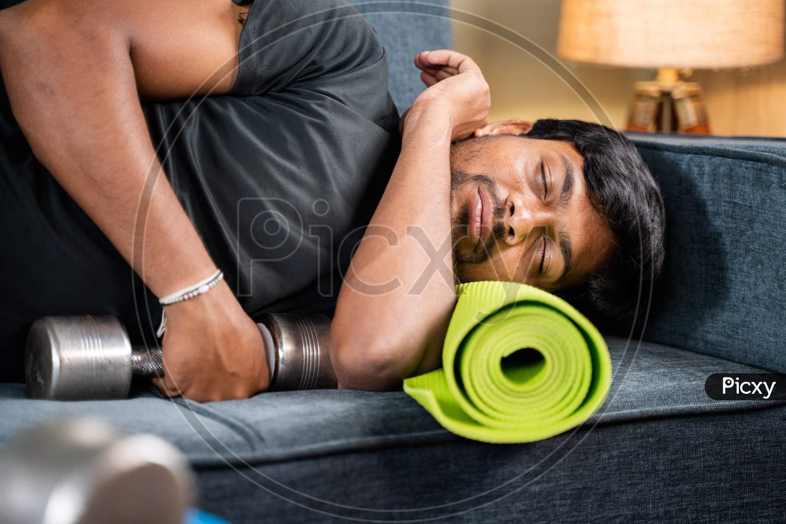 Tired Young Man Sleeping On Sofa By Holding Dumbbells And Yoga Mat - Concept Of Healthcare, Lifestyle And Fitness Man Resting After Workout In Home Gym.