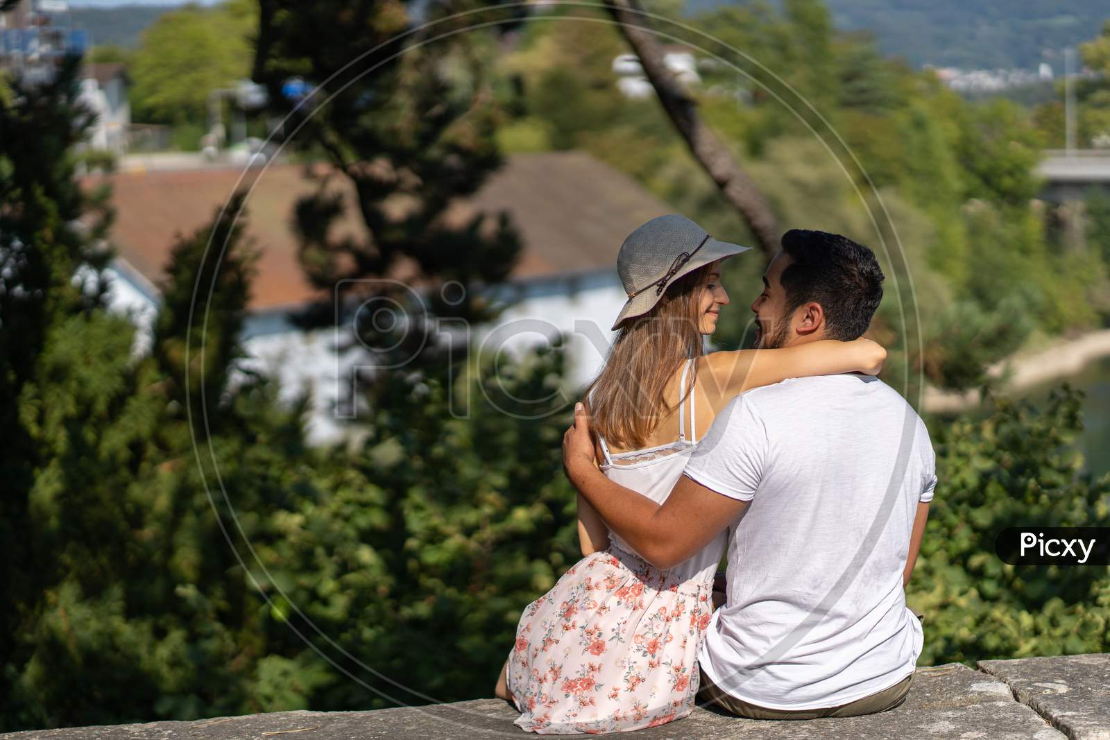 Young couple Sitting On Stone Wall Embracing, Woman With Straw Hat Has A Smile On Face.