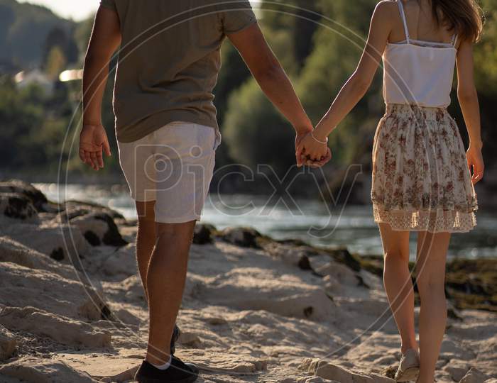 Rear view of Young Couple, Walking With Clasped Hands In Sunshine On Riverside At Late Afternoon In September.