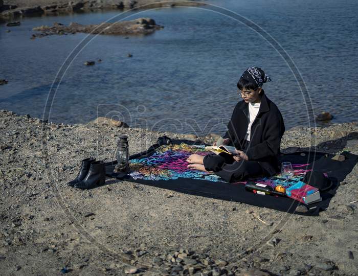 Young Beautiful Indian Girl Reading A Book Sitting On A Blanket In The Mountains With A Lake Nearby. Concept Of Having Picnic In A City Park During Winter Holidays Or Weekends.