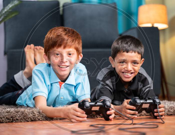 Two Multi Ethnic Kids Busy In Playing Videogame Using Gamepad While Lying On Floor At Home - Concept Of Children Unhealthy Playing Position And New Technology Addiction