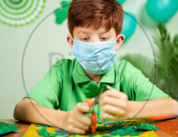 Young Kid Busy In Preparing Shamrock Leaves For Saint Patricks Day Festival Celebrations At Home With Face Mask During Coronavirus Covid-19 Pandemic.