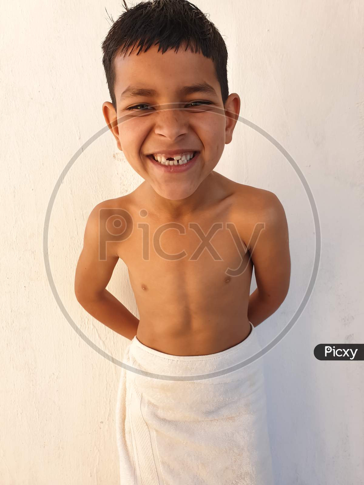 Studio shot of a shirtless little boy with funny big smile pose, A Indian kid showing his broken teeth with crazy smile, A small boy with wearing white towel and looking at camera.