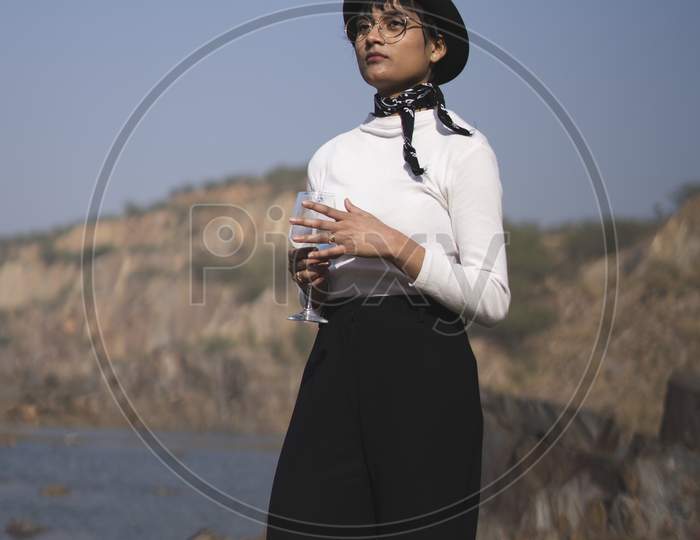 Long Shot Of An Indian Girl Standing Near A Lake Surrounded With Mountains, Wearing White Top And Black Pants.