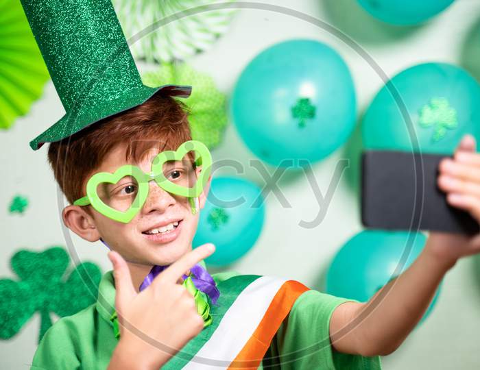 Young Kid On Video Call During Saint Patricks Day Celebration On Mobile Phone And Showing Decorated Background Over Video Call - Concept Of Distant Festivels Due To Coronavirus Or Covid-19 Pandemic.