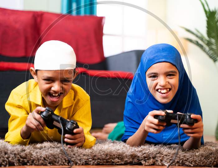 Two Muslim Sibling Kids Playing Videogame Using Joystick While Lying On Floor At Home - Concept Of Children Unhealthy Playing Position And Gaming Addiction