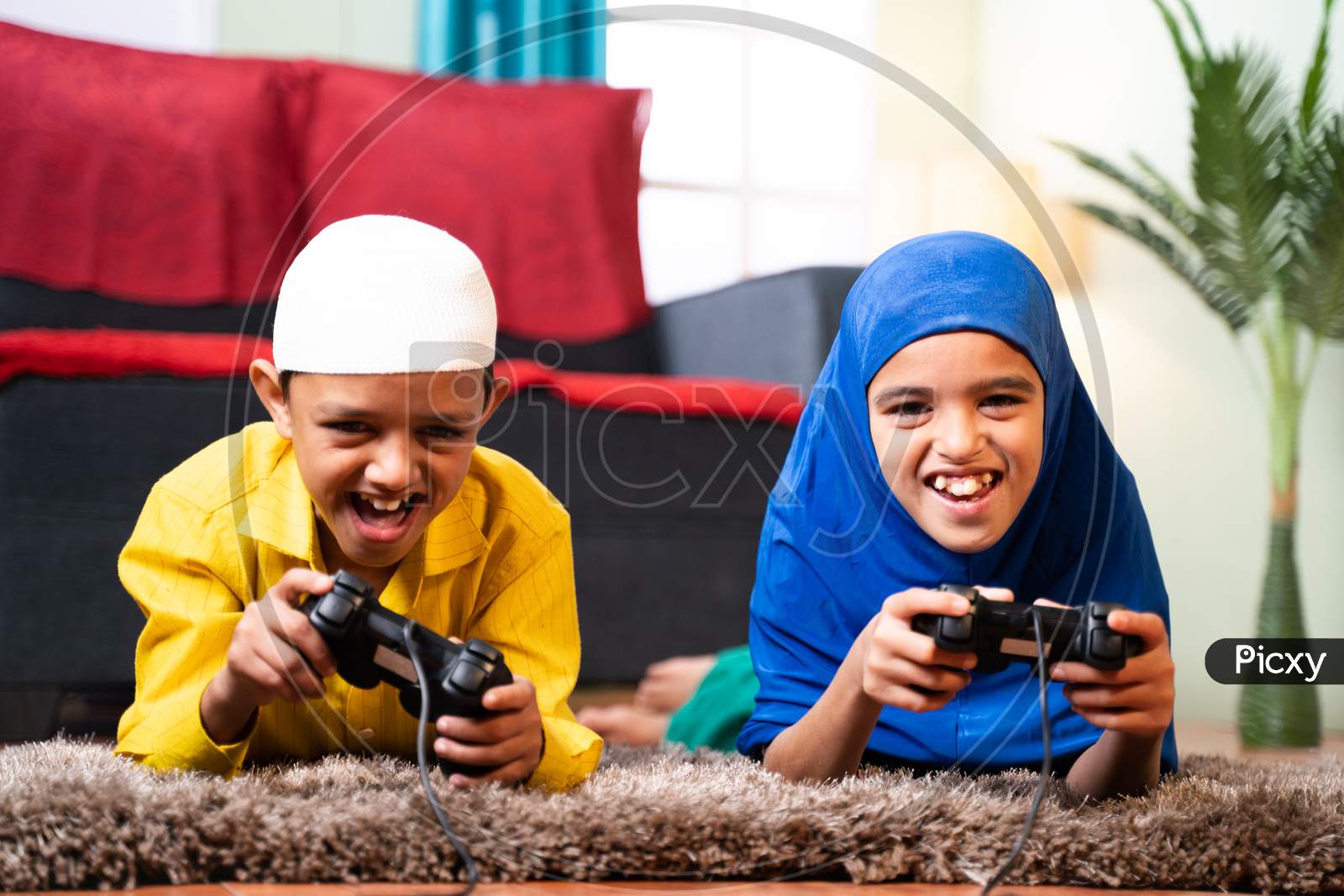 Two Muslim Sibling Kids Playing Videogame Using Joystick While Lying On Floor At Home - Concept Of Children Unhealthy Playing Position And Gaming Addiction