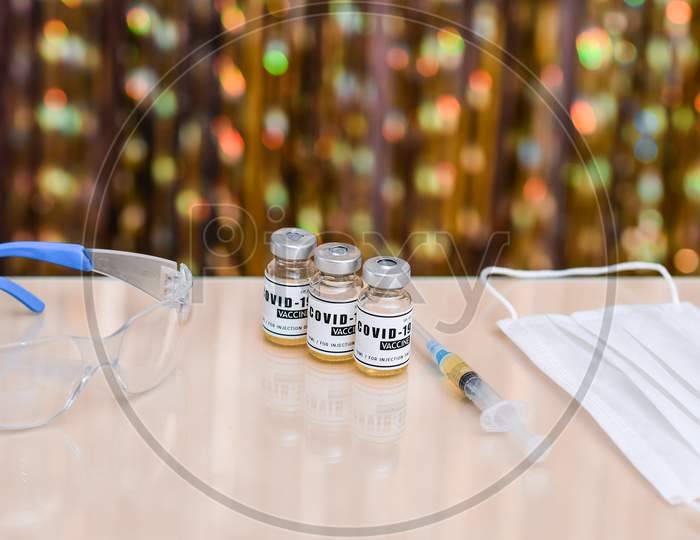 Coronavirus Vaccine Several Ampoules And Syringe Injection, Safety Glasses, Mask. Treatment From Corona Virus Infection. On A Golden Bokeh Background.