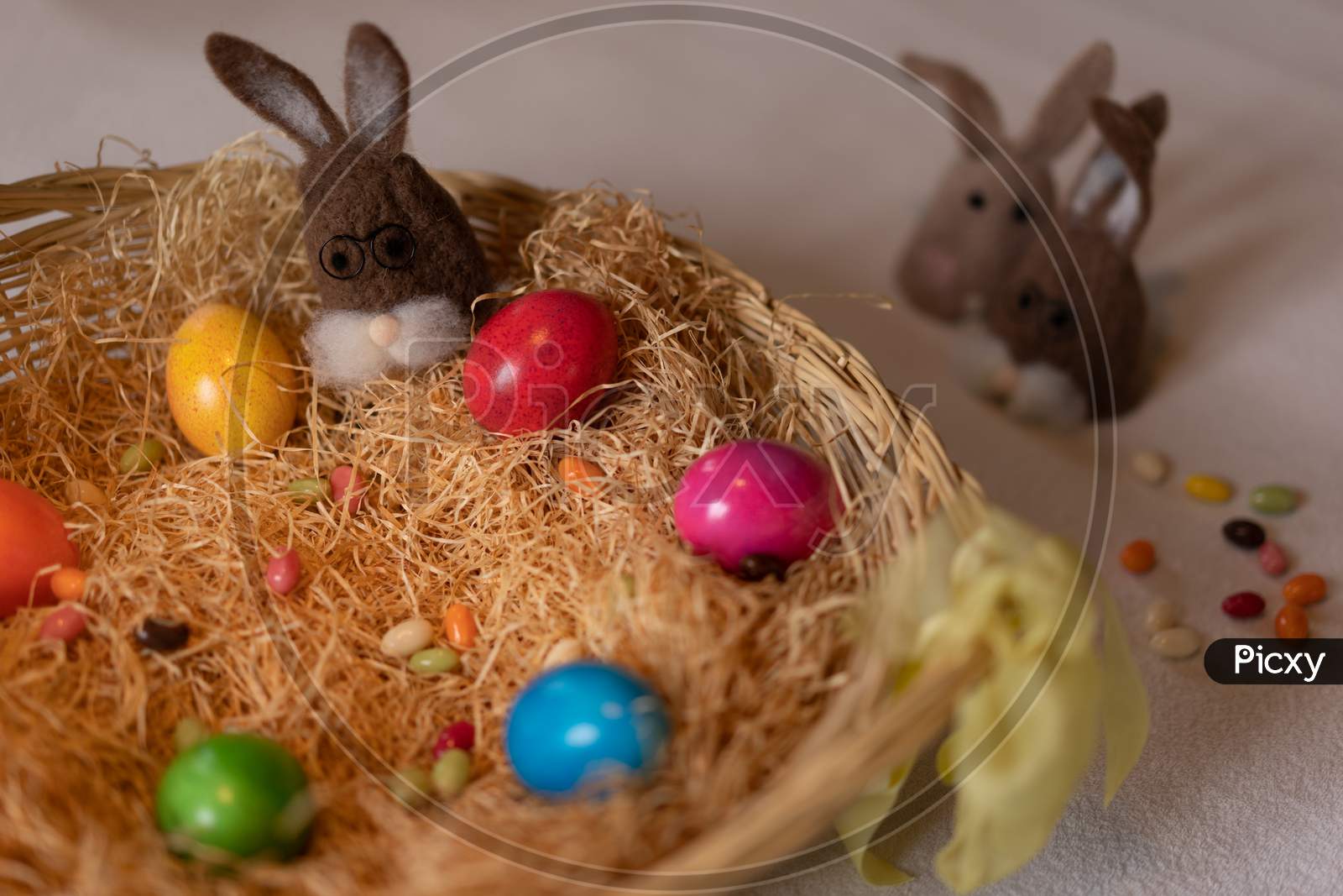 Colored Easter Eggs And Caps Of Brown Felt Bunnies In Nest Of Straw With Colorful Sweet Sugar Eggs. Felt Bunnies To Keep Eggs Warm.