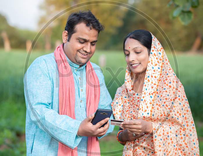 Happy Indian Rural Farmer Couple Using Smartphone To Make Online Payment With Debit Card In Agricultural Field, Shopping On Internet With Cellphone Secure Banking Service System Concept.