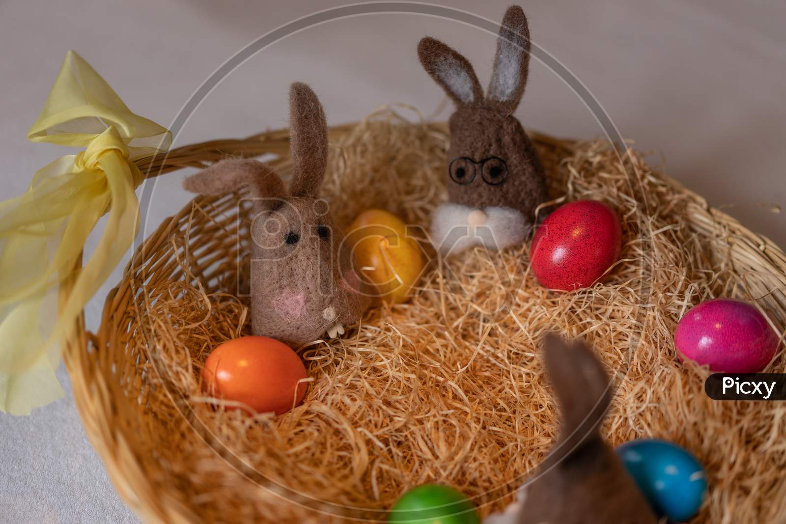 Brown Felt Bunnies With Multicolored Easter Eggs In Nest Of Straw With Colorful Sweet Sugar Eggs.