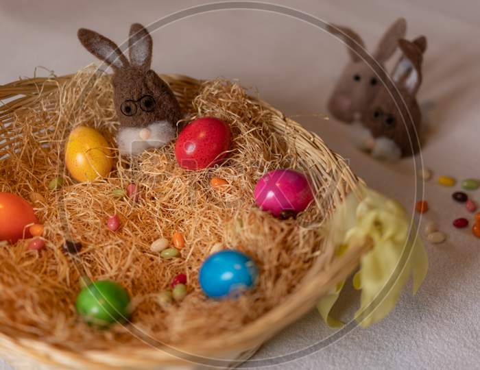 Colored Easter Eggs And One Brown Felt Bunnies In And Two Separated Other Out Of The Nest Of Straw With Colorful Sweet Sugar Eggs.