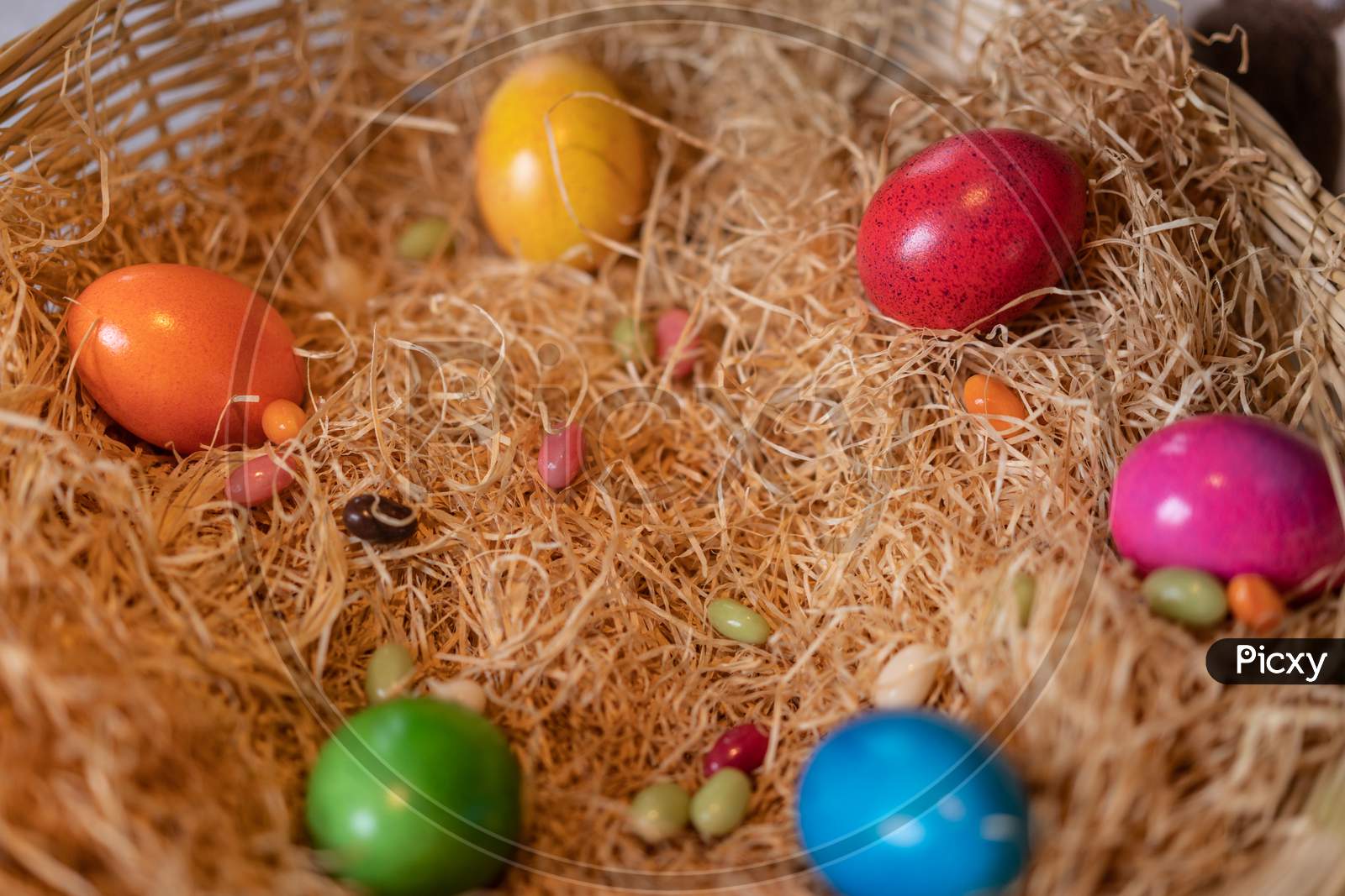 Yellow, Red, Green, Blue, Pink And Orange Colored Cooked Easter Eggs Put In A Nest Of Straw In Straw Basket On White Ground.
