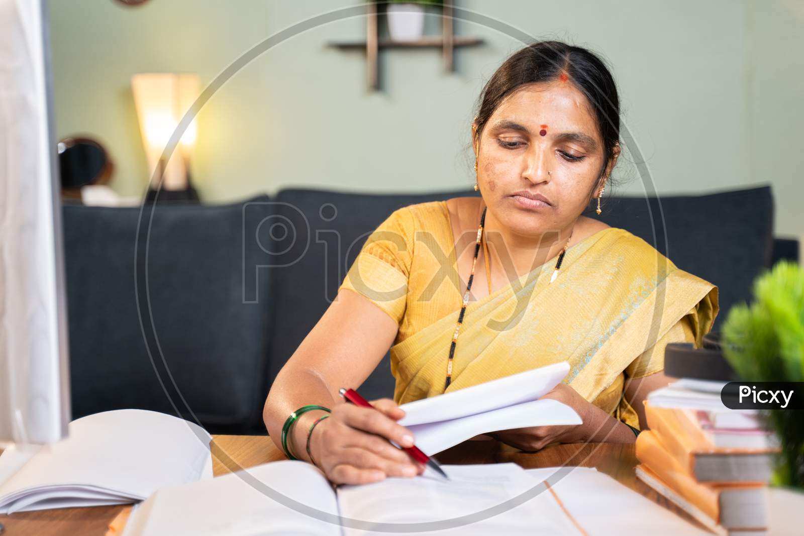 Indian Woman Professor Correcting Answer Sheets Of Books By Referring Books At Home At Working Desk.