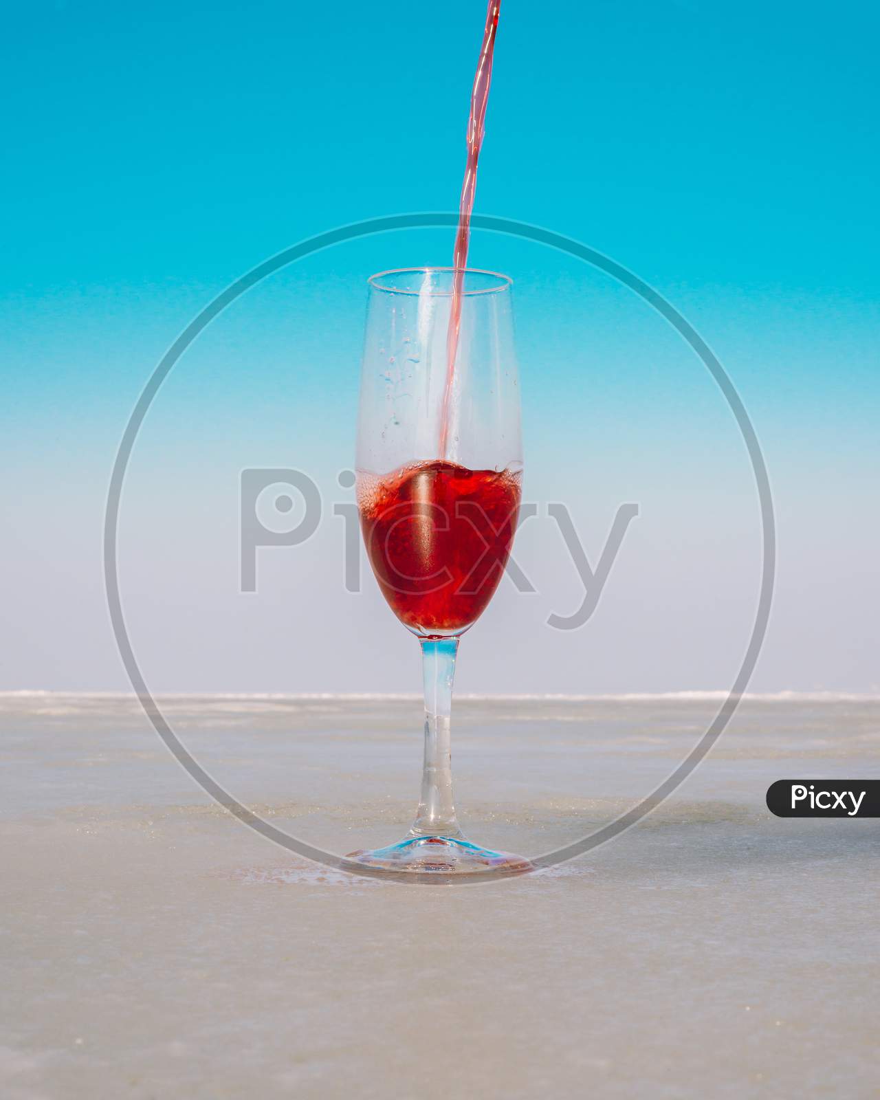 A stream of red wine is poured into a transparent glass against a blue sky on a sea coast