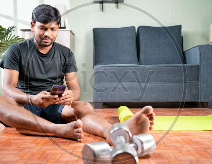 Wide Angle Shot Of Young Man Busy Using Mobile Phone During Work Out At Home - Millennial Checking Online Exercise Tutorials For Workout