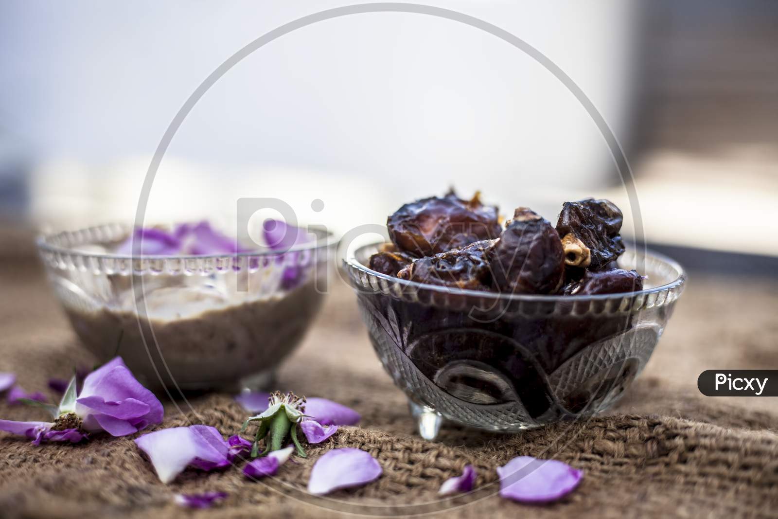 Close Up Shot Of Popular Indian & Asian Winter Dessert In A Transparent Glass Bowl I.E. Khajor Ka Halwa Or Dates Confection With Raw Dates And Some Rose Petals On Brown Colored Surface.