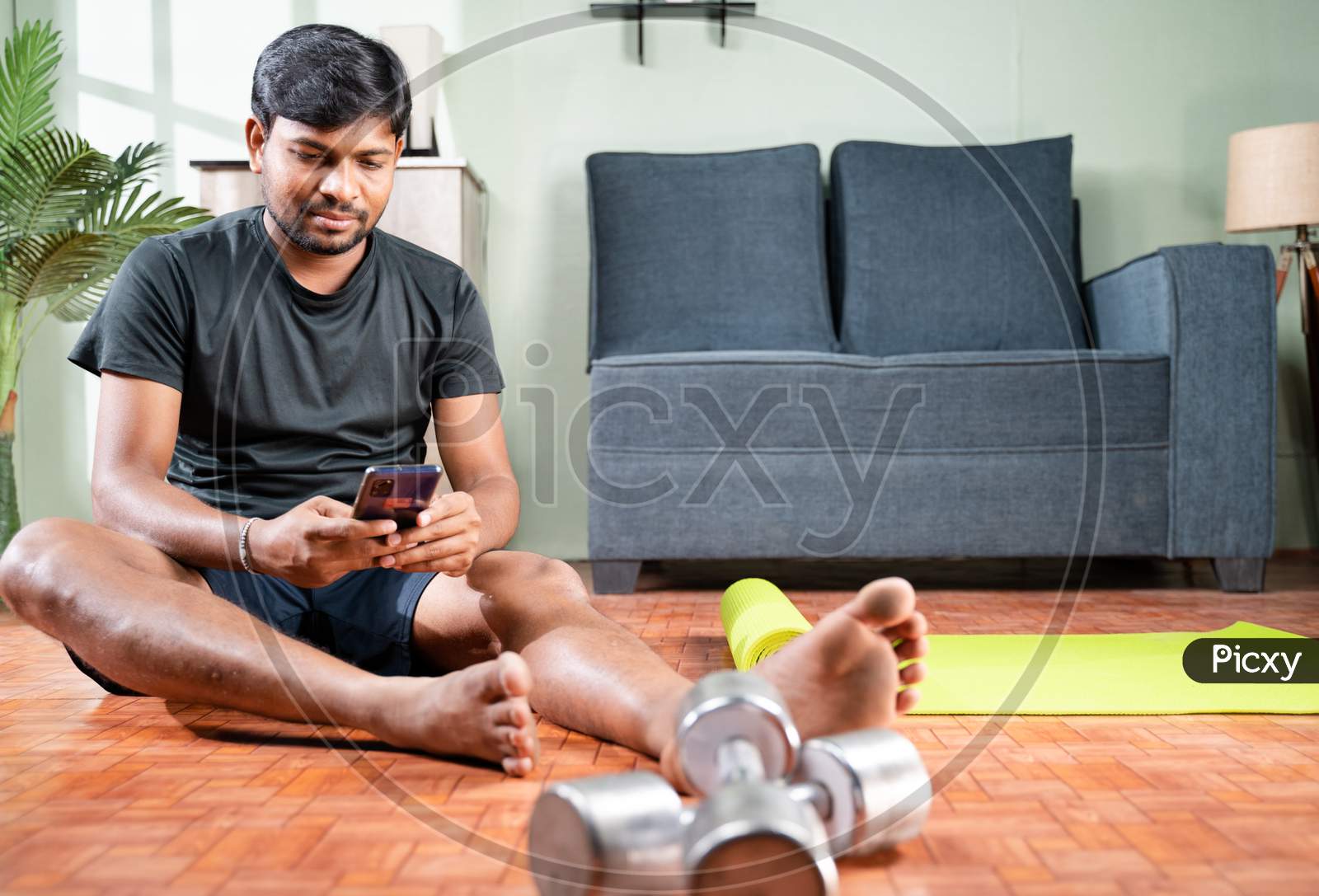 Wide Angle Shot Of Young Man Busy Using Mobile Phone During Work Out At Home - Millennial Checking Online Exercise Tutorials For Workout
