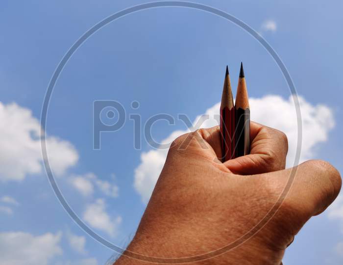 South Indian Male Hand Holding Two Sharp Pencils. Cloud Background