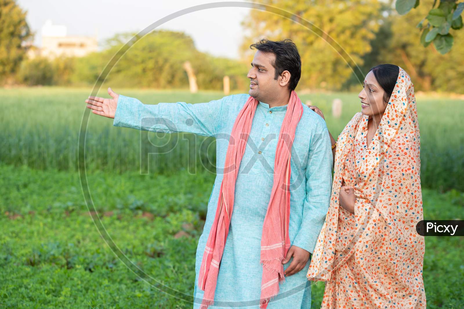 Happy Young Indian Rural Farmer Couple In Agricultural Field Looking At Each Other Laughing. Copy Space To Write Text. Man Wearing Kurta And Woman Wearing Saree,