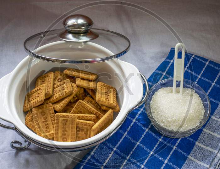 Indian Famous Brown Glucose Biscuit Kept Inside White Glass Bowl And White Sugar Container Kept Beside Bowl Of Biscuit.