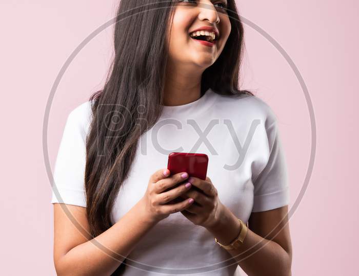 Smiling Indian Young Woman Wears White T Shirt Using Smartphone Or Cellphone Against Pink Background
