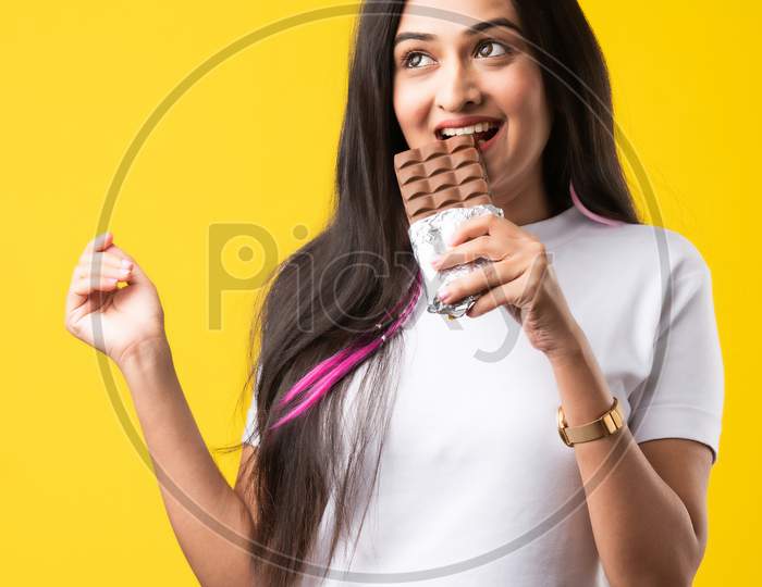 Happy & Pretty Indian Asian Young Woman Standing Isolated Over A Yellow Background Eating Chocolate