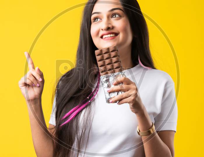 Happy & Pretty Indian Asian Young Woman Standing Isolated Over A Yellow Background Eating Chocolate
