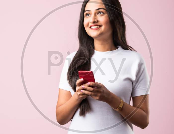 Smiling Indian Young Woman Wears White T Shirt Using Smartphone Or Cellphone Against Pink Background