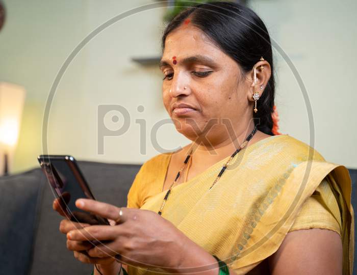 Serious Indian Woman Busy Using Mobile Phone While Sitting On Sofa At Home - Concept Of Social Media Bullying And Online Messaging.