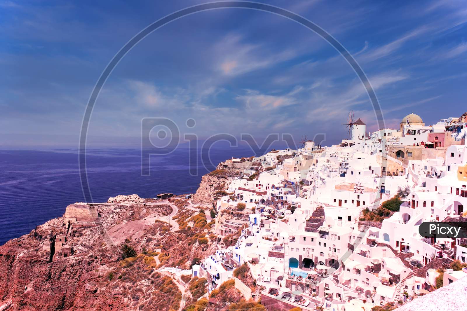 Santorini, Greece - September 11, 2017: Wide Angle Panoramic View Of Oia Santorini White Buildings On The Hillside Facing North Against The Blue Sky. Cityscape Of Famous Greek Island