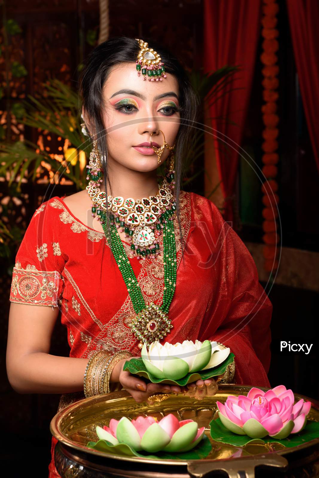 Portrait Of Very Beautiful Young Indian Bride In Luxurious Bridal Costume With Makeup And Heavy Jewellery Holding A Lotus In Studio Lighting Indoor. Lifestyle Wedding Fashion.
