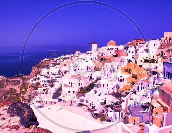 Santorini, Greece: Beautiful City Of Oia ( Ia ) On A Hill Of White Houses With Blue Roof And Windmills Against Dramatic Pink Sky, Located In Greek Cyclades Islands In Mediterranean Sea