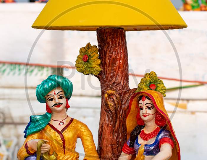 Beautiful Handmade Dolls Of Miniature Villagers With A Decorative Table Lamp Are Displayed In A Shop For Sale In Blurred Background. Indian Handicraft.
