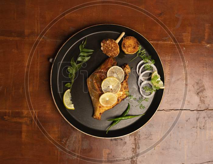 Pomfret Fish Fry- Deeply Fried Two Pomfret Fishes Which Is Well Garnished With Lemon And Onion In A White Ceramic Plate With Wooden Background