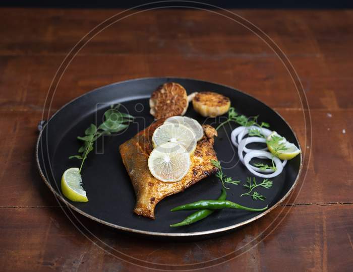 Pomfret Fish Fry- Deeply Fried Two Pomfret Fishes Which Is Well Garnished With Lemon And Onion In A White Ceramic Plate With Wooden Background