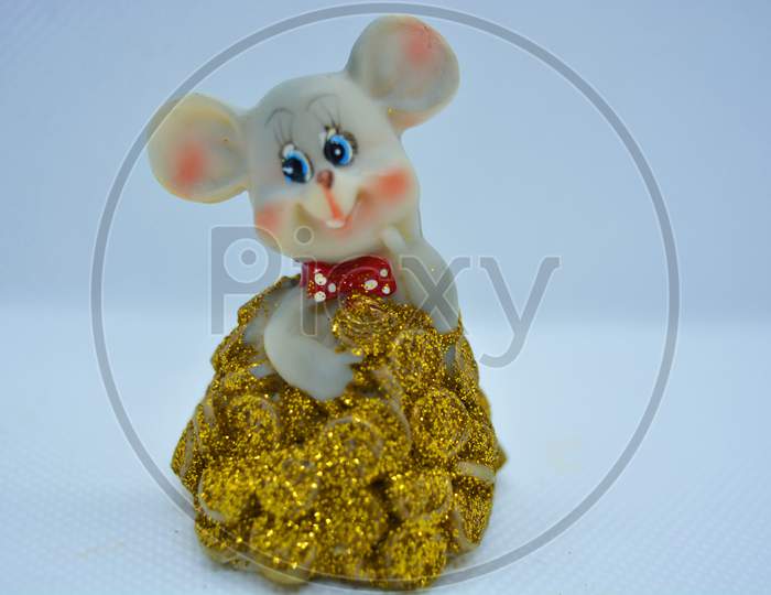 A figurine of a cute gray mouse peeking out of a mountain of yellow coins is located on a white fabric background.