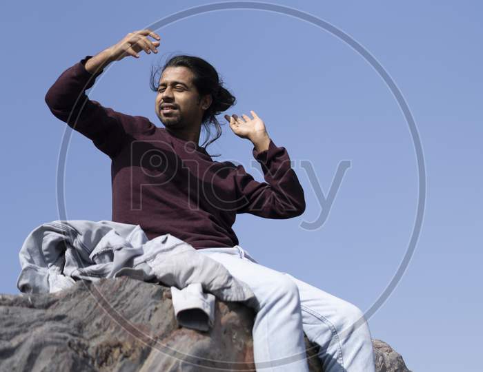 Young Indian Boy Standing On The Top Of Cliff Wearing A Snow Jacket And Track Pants. Enjoying And Freedom Concept.