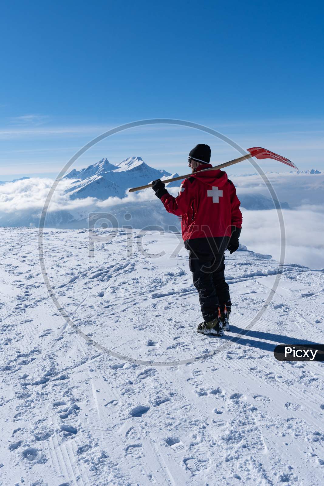 Ski patroller On Snow Caped Mountain Is Walking Against Sun With A Red Rescue Jacket And A Shovel On His Shoulders.