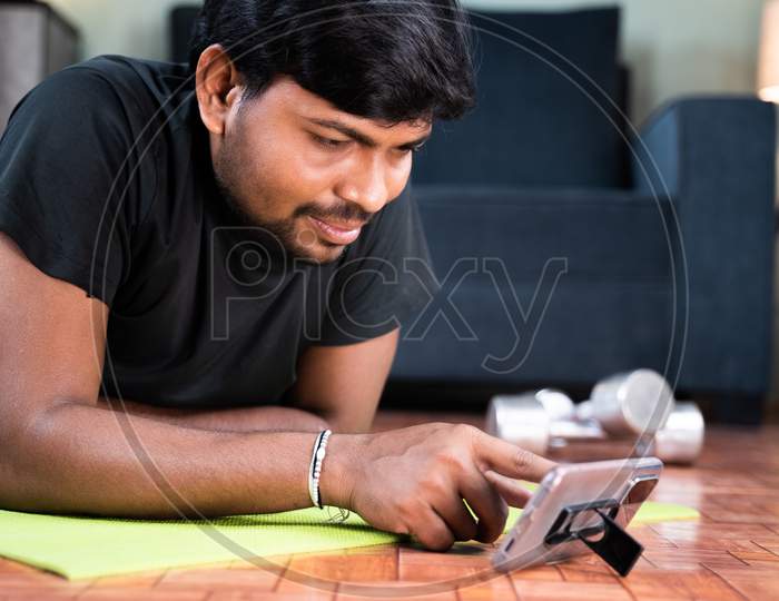 Young Man On Yoga Mat Checking Online Workout Or Yoga Tutorials On Mobile Phone At Home - Concept Of New Normal, Home Gym Due To Coronavirus Covid-19 Pandemic Lockdown.