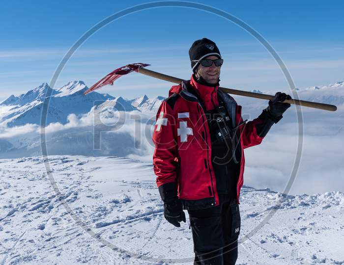 Ski Patroller On Snow Caped Mountain Is Standing Against Sun With A Red Rescue Jacket And A Shovel On His Shoulders
